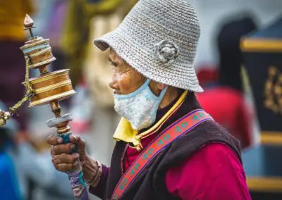 An elderly Tibetan or Himalayan woman holds a traditional prayer wheel, her face focused and contemplative. She wears a flower-adorned woven hat, maroon top, yellow scarf, and a strap with Tibetan script. The ornate prayer wheel, with brass tones and bells, reflects her deep spiritual devotion.
