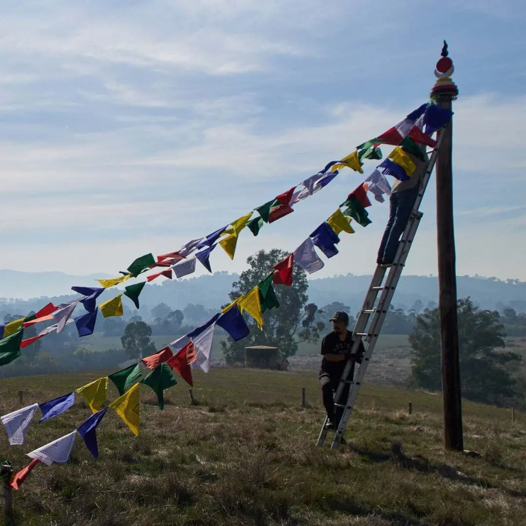 flag-raising volunteers raising tibetan prayer flags on Saka Dawa at Land of Shambhala. One is on a ladder and the other is helping the person connect the flags to the pole that has a sun moon rahu and kalgni disk on it with a nada rising to the sky.