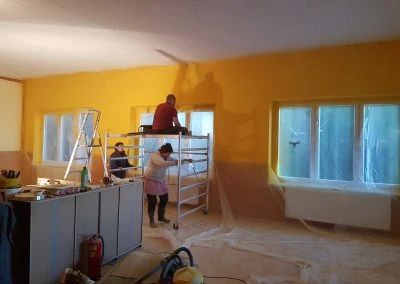 A group of volunteers is renovating the colorful Kalapa Tibetan Buddhist center. The space has bold yellow walls, with a man and two women actively painting using various tools and workbenches. The room contains construction materials, a ladder, and floor coverings for protection.