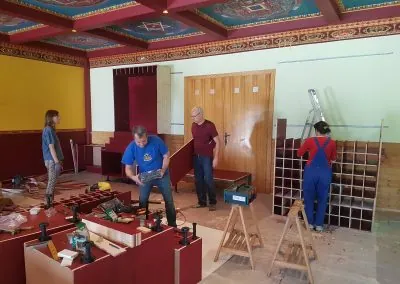 A group of volunteers is renovating the vibrant Kalapa Tibetan Buddhist Gompa. The space features bold yellow and deep red walls, with an intricately decorated ceiling. Two men and two women are assembling wooden furniture, using various tools and workbenches. The room is filled with construction materials, a ladder, and protective floor covering.